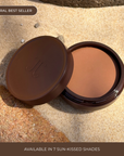 A compact cream bronzer described as a 'VIRAL BEST SELLER' with text indicating it is available in 7 sun-kissed shades, displayed on a sandy surface.