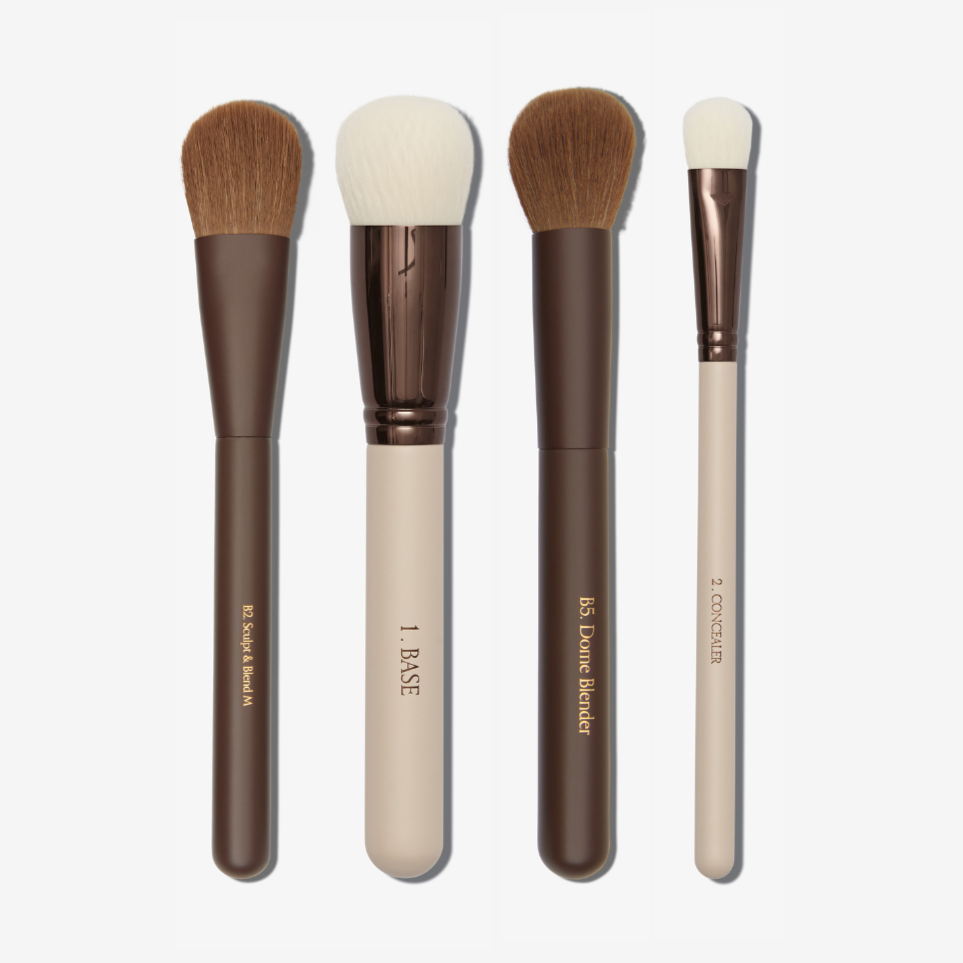A collection of four makeup brushes including a blending brush, a base brush, a dome blender brush, and a concealer brush.