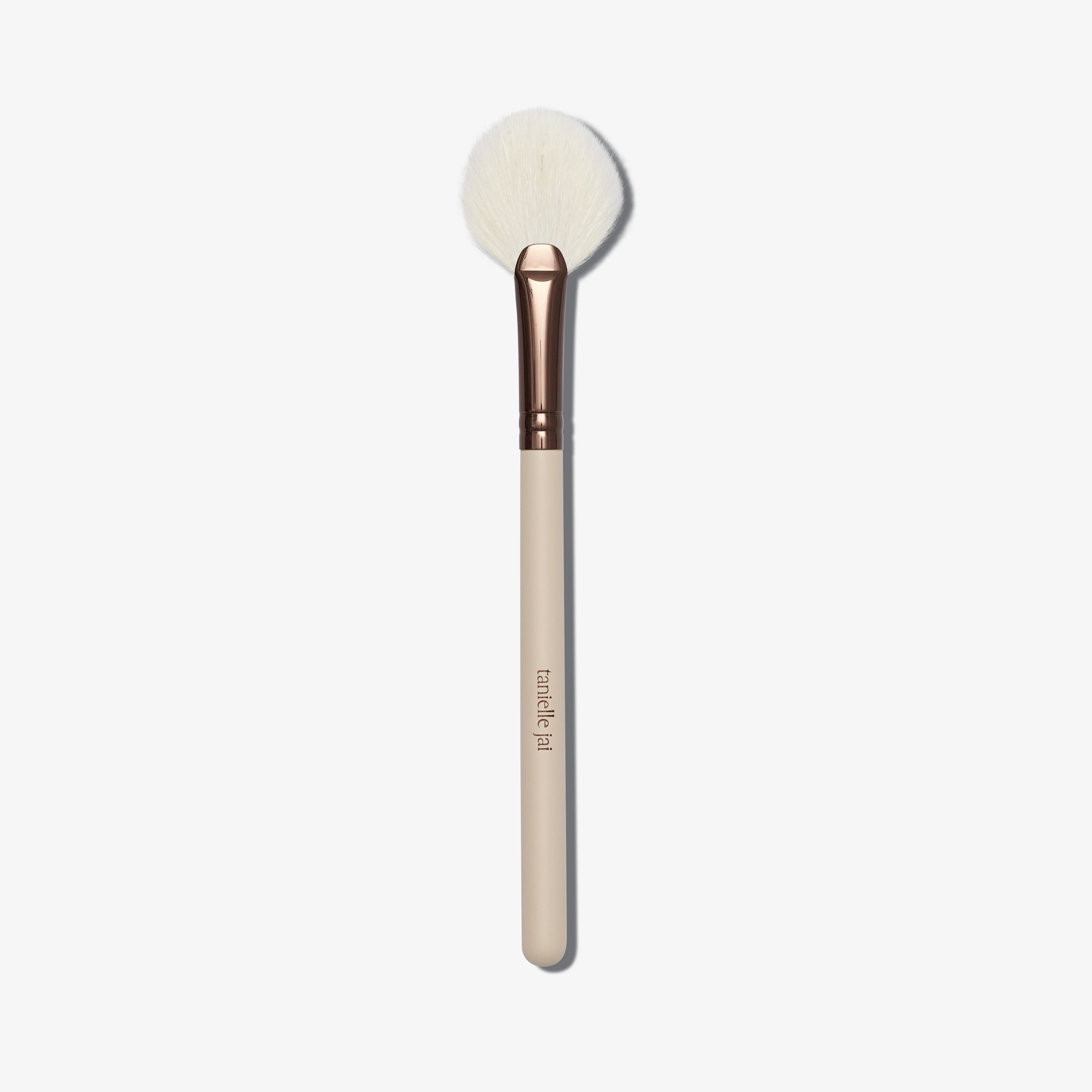 A cream coloured fan shaped makeup brush with white brush hairs with logo 'taniellejai'